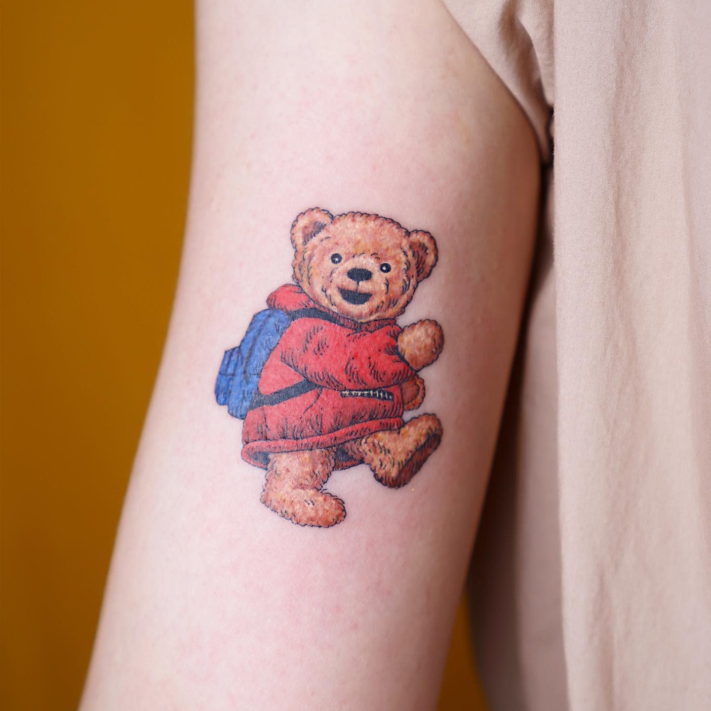 Tattoo of a smiling teddy bear wearing a red hoodie and blue backpack on someone's arm